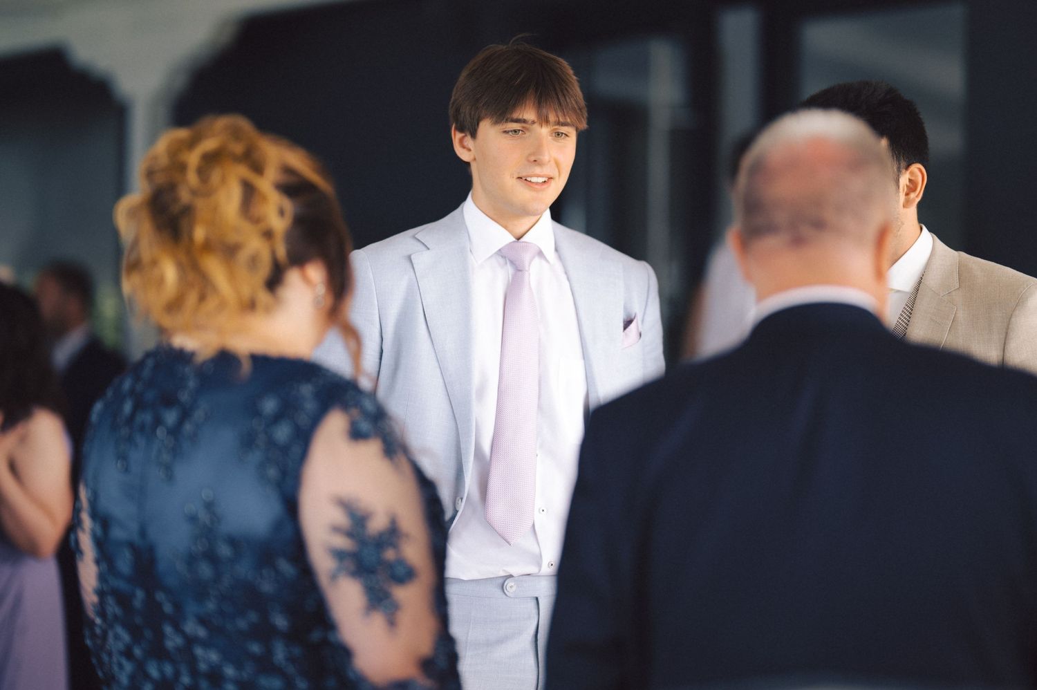 Someone in a light gray suit and pink tie stands between formal event attendees in a social gathering setting.