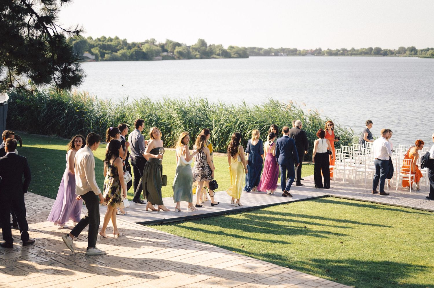 A wedding party lines up for photos along a serene lakeside with a grassy lawn and scenic water view.