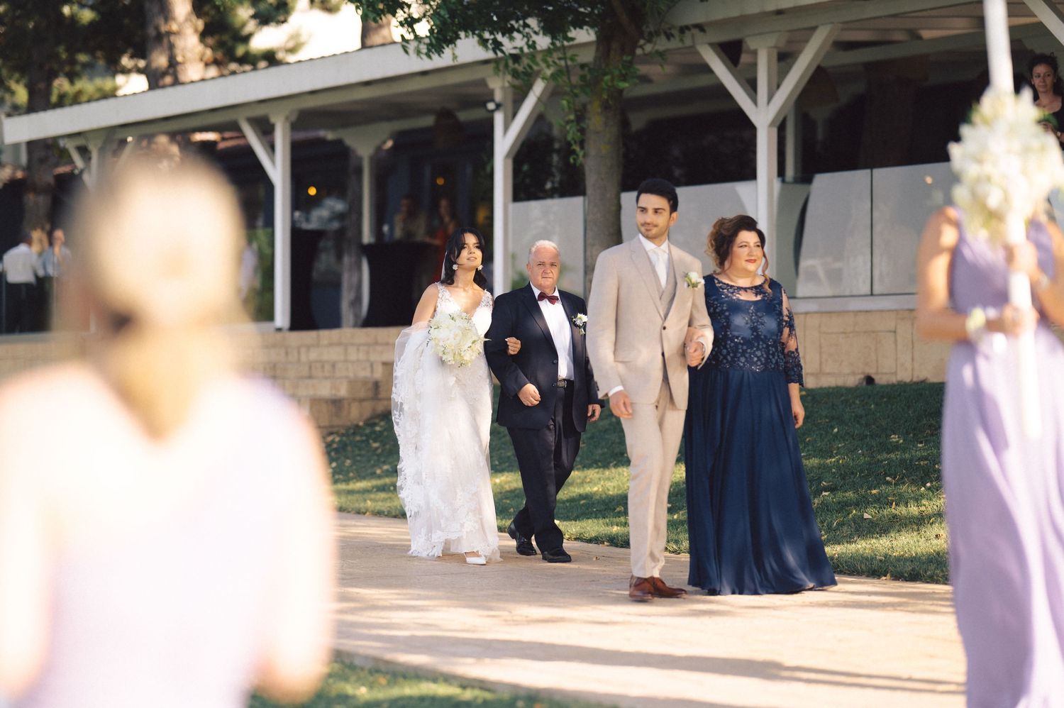 People in formal attire including elegant white and navy blue gowns gather outdoors at a special event.