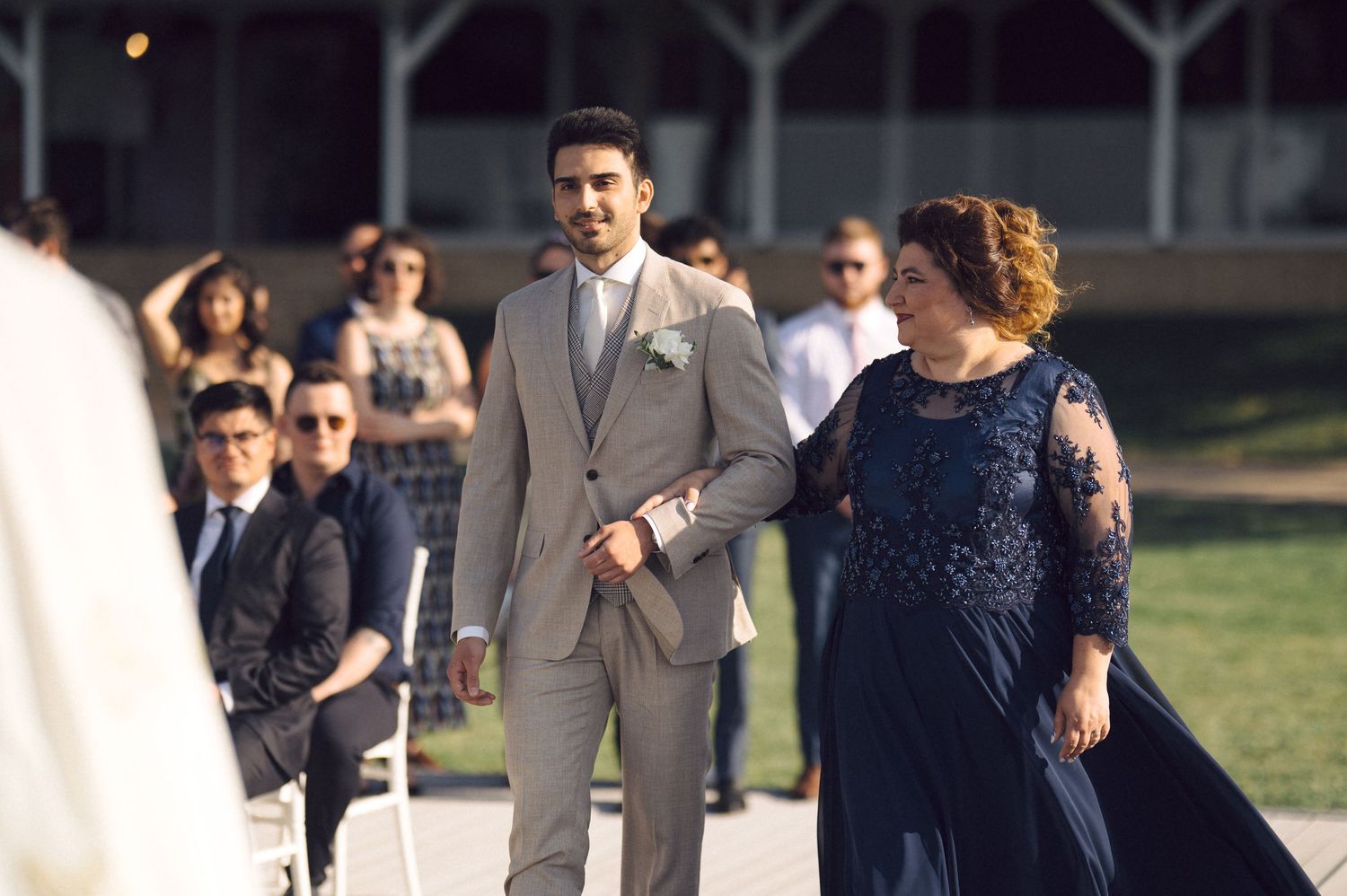 A groom in a light gray suit walks alongside someone in a navy blue lace dress during an outdoor wedding ceremony.