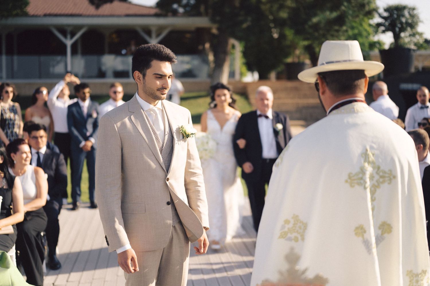 A person in a beige suit facing a religious officiant in white robes during an outdoor ceremony.