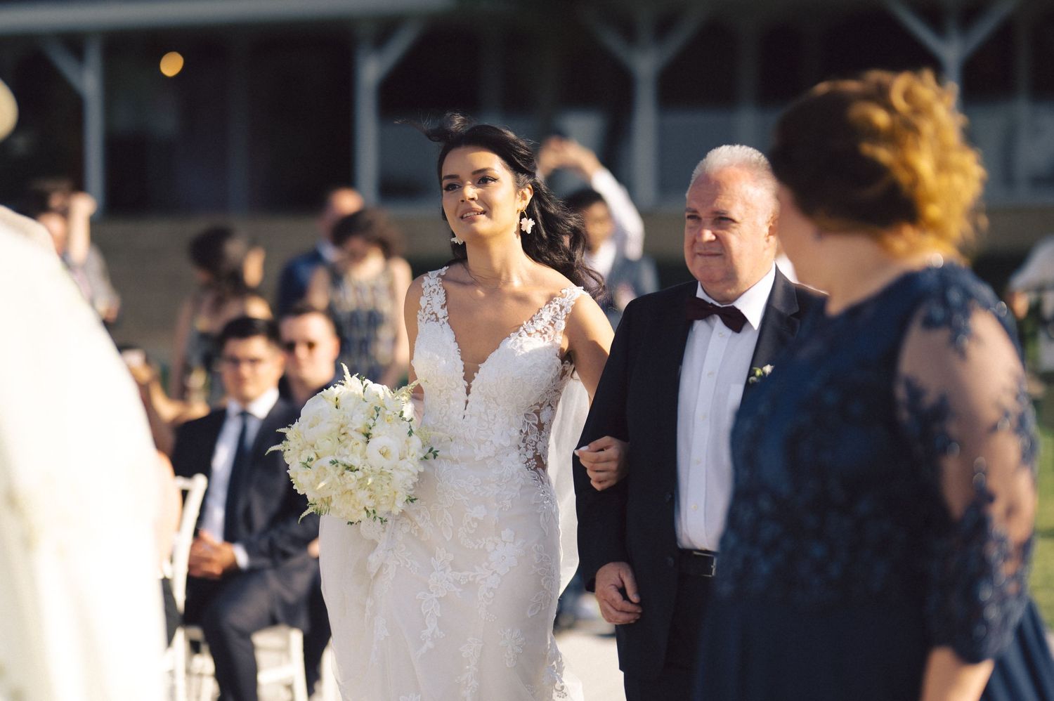 At a formal outdoor wedding ceremony, a bride in an elegant white lace gown walks down the aisle with her escort.
