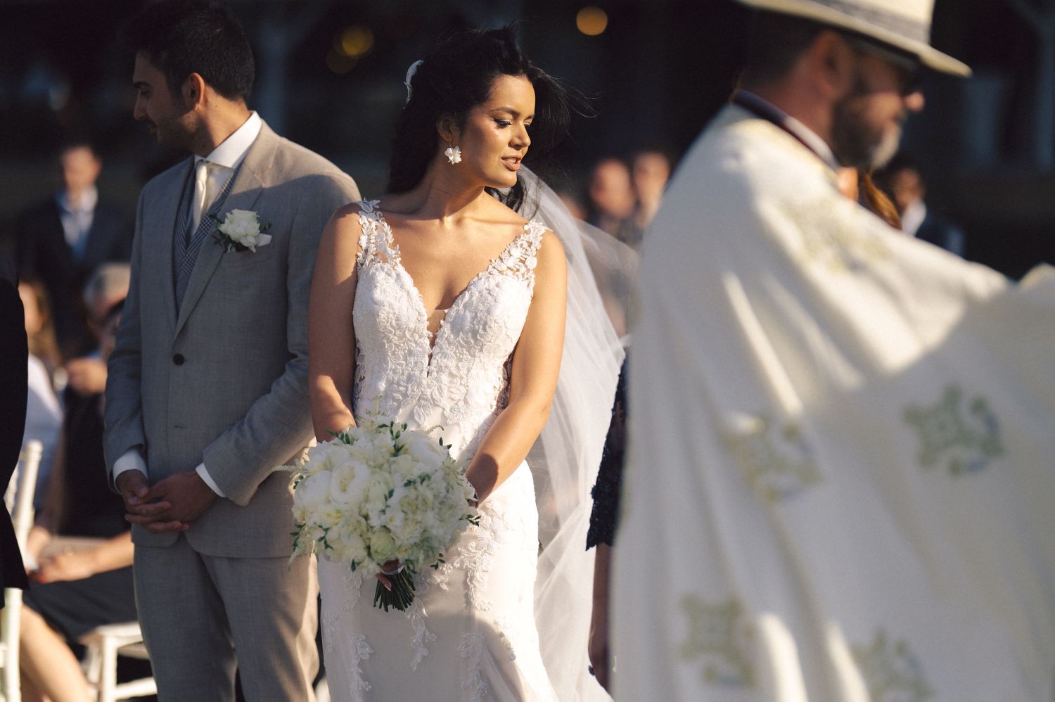 A bride in an elegant lace wedding gown holds a soft blue-green bouquet during an outdoor ceremony.