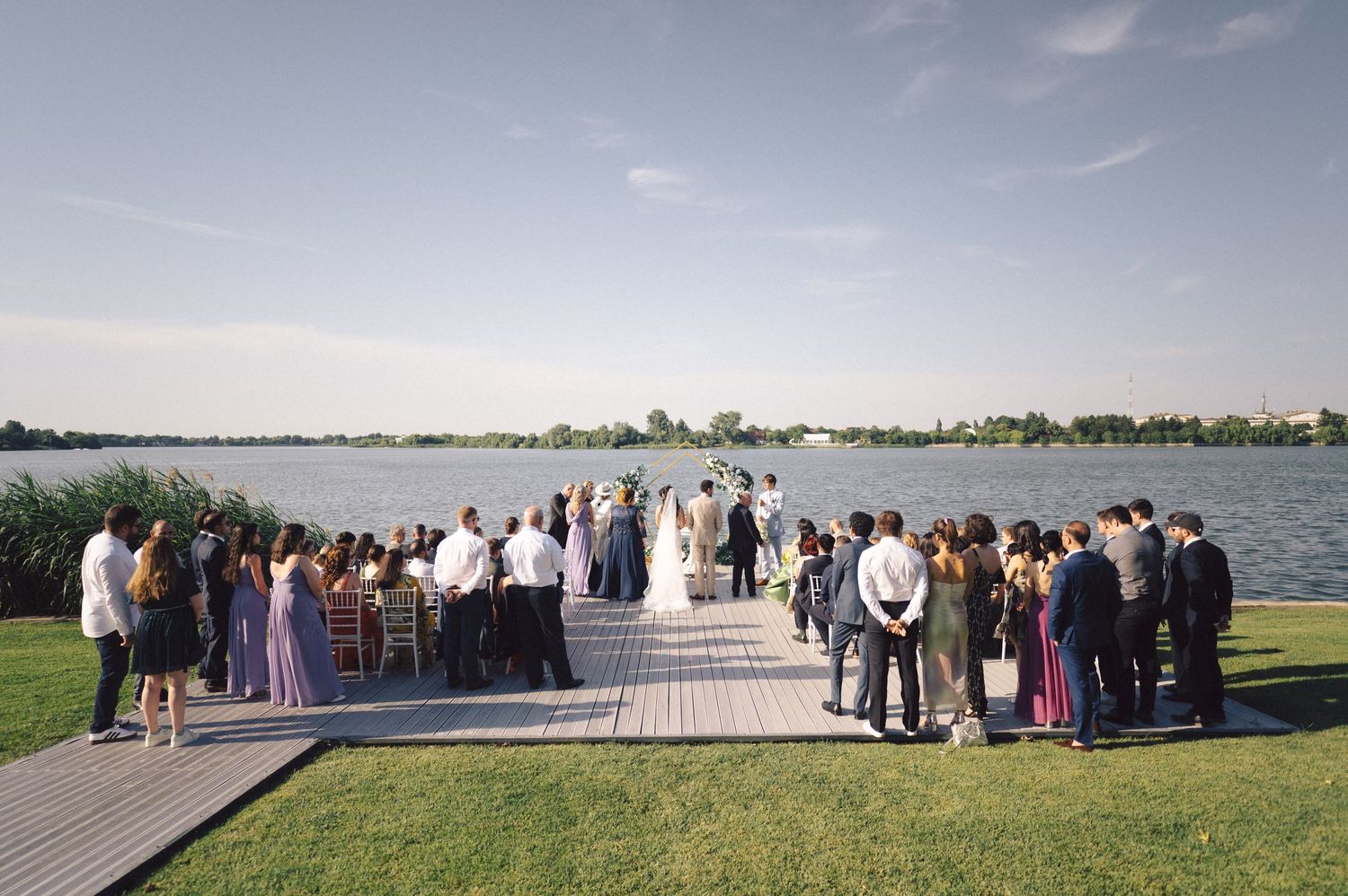 A wedding ceremony takes place on a waterfront dock with guests gathered on both sides and a lake view in the background.