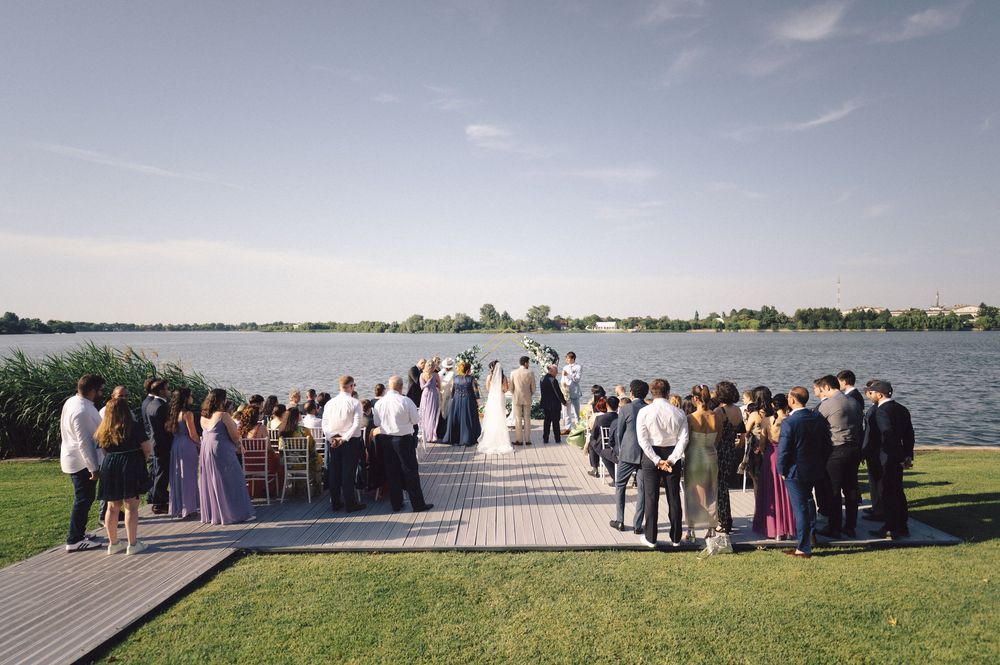 A wedding ceremony takes place on a waterfront dock with guests gathered on both sides and a lake view in the background.