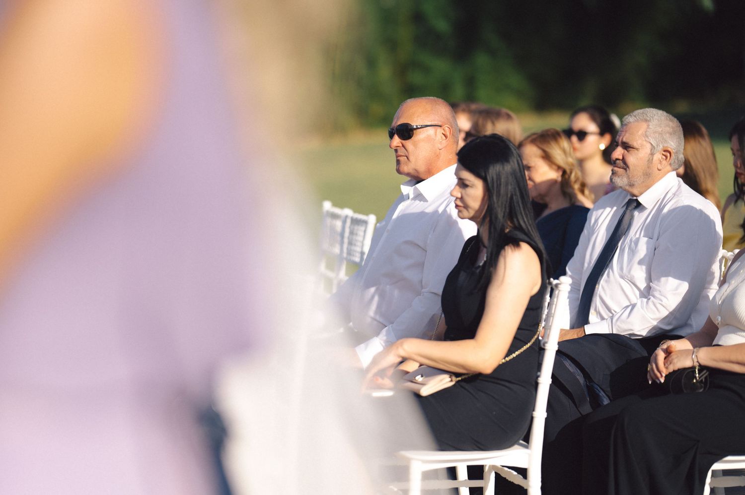 Wedding guests sit together at outdoor tables during a daytime ceremony in a garden setting.
