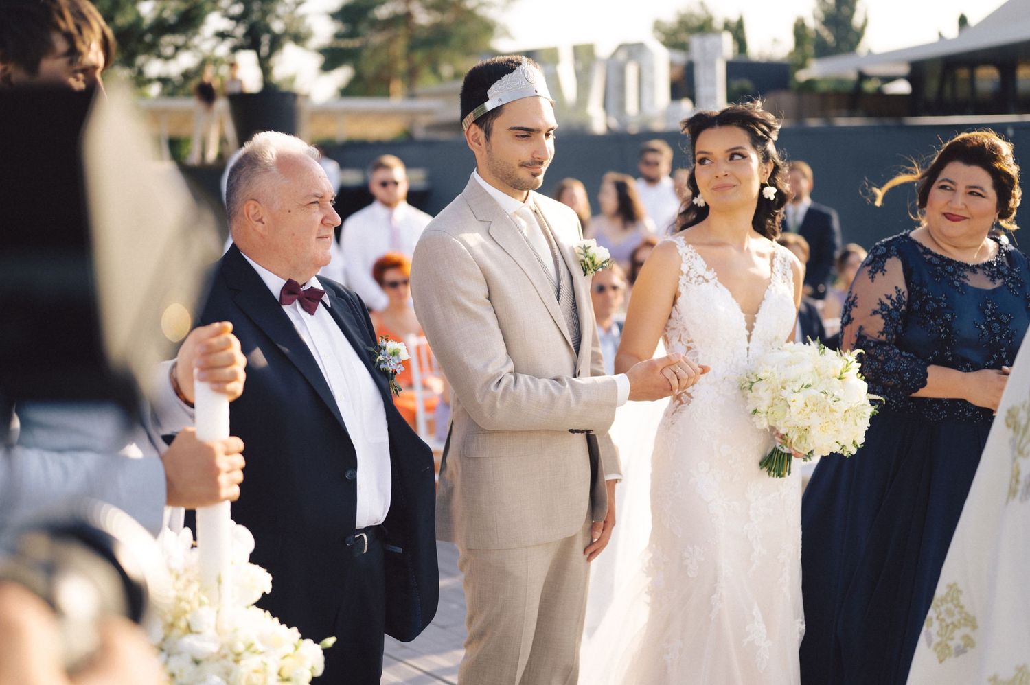 Wedding party members gathered outdoors on a sunny day with the bride holding a white bouquet.