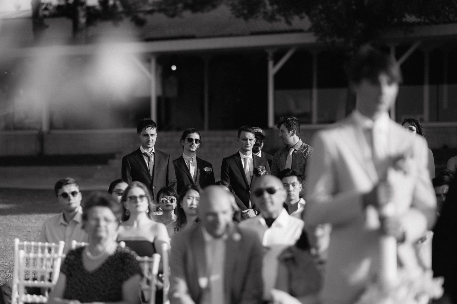A black and white vintage photograph shows a group of people seated outdoors wearing sunglasses and formal attire.