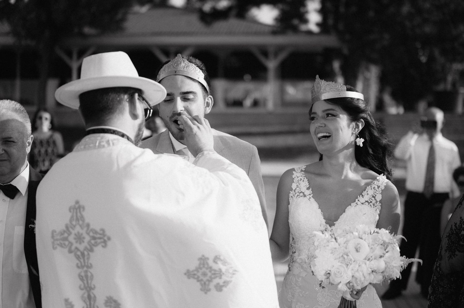 Black and white photo of an Orthodox priest performing a wedding ceremony with a smiling bride holding white flowers.