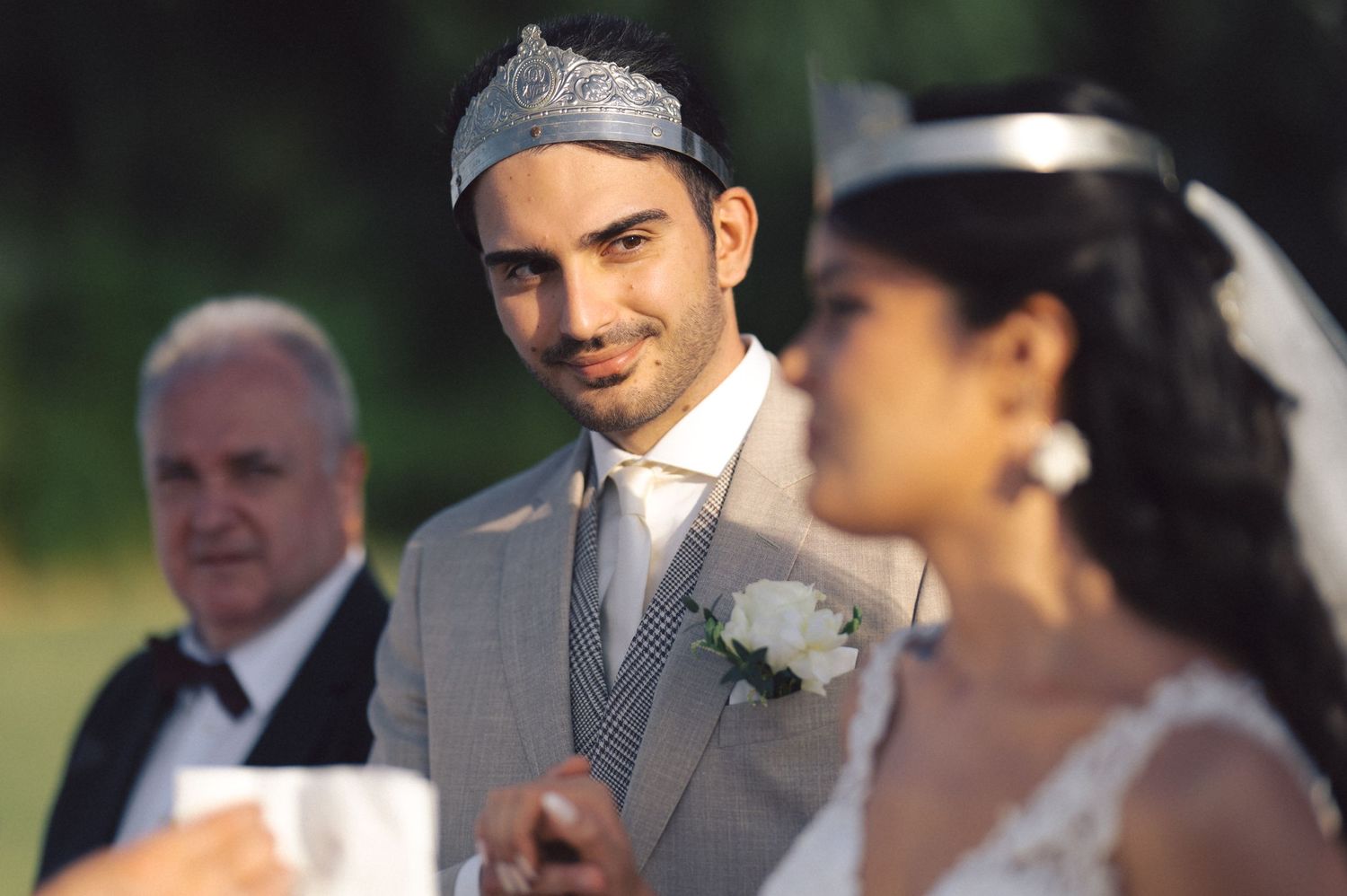 A joyful outdoor wedding ceremony with the groom in a light gray suit and silver tie, smiling during the vows.