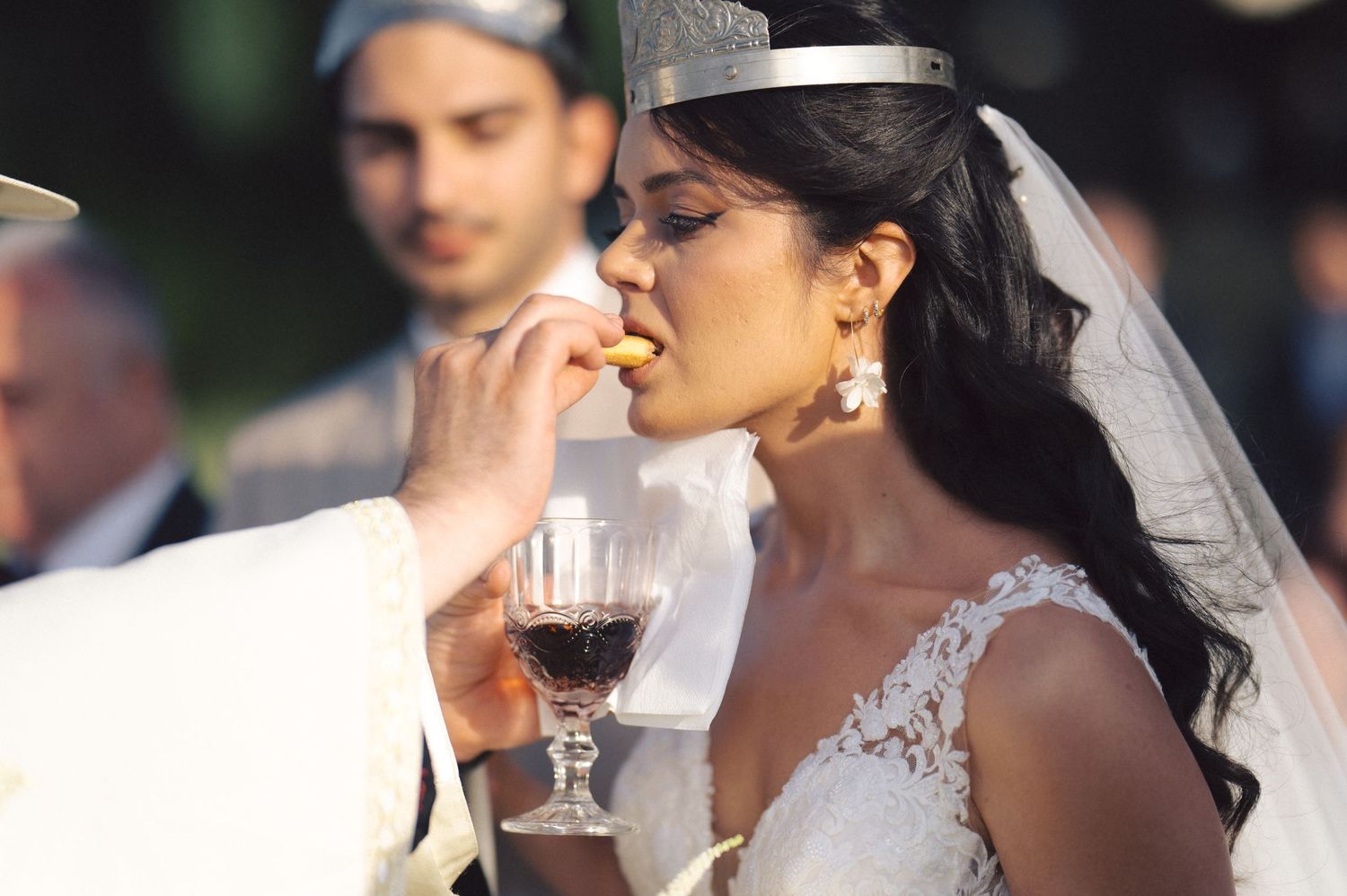 A bride in a white lace wedding dress enjoys a sip of red wine during an outdoor celebration.