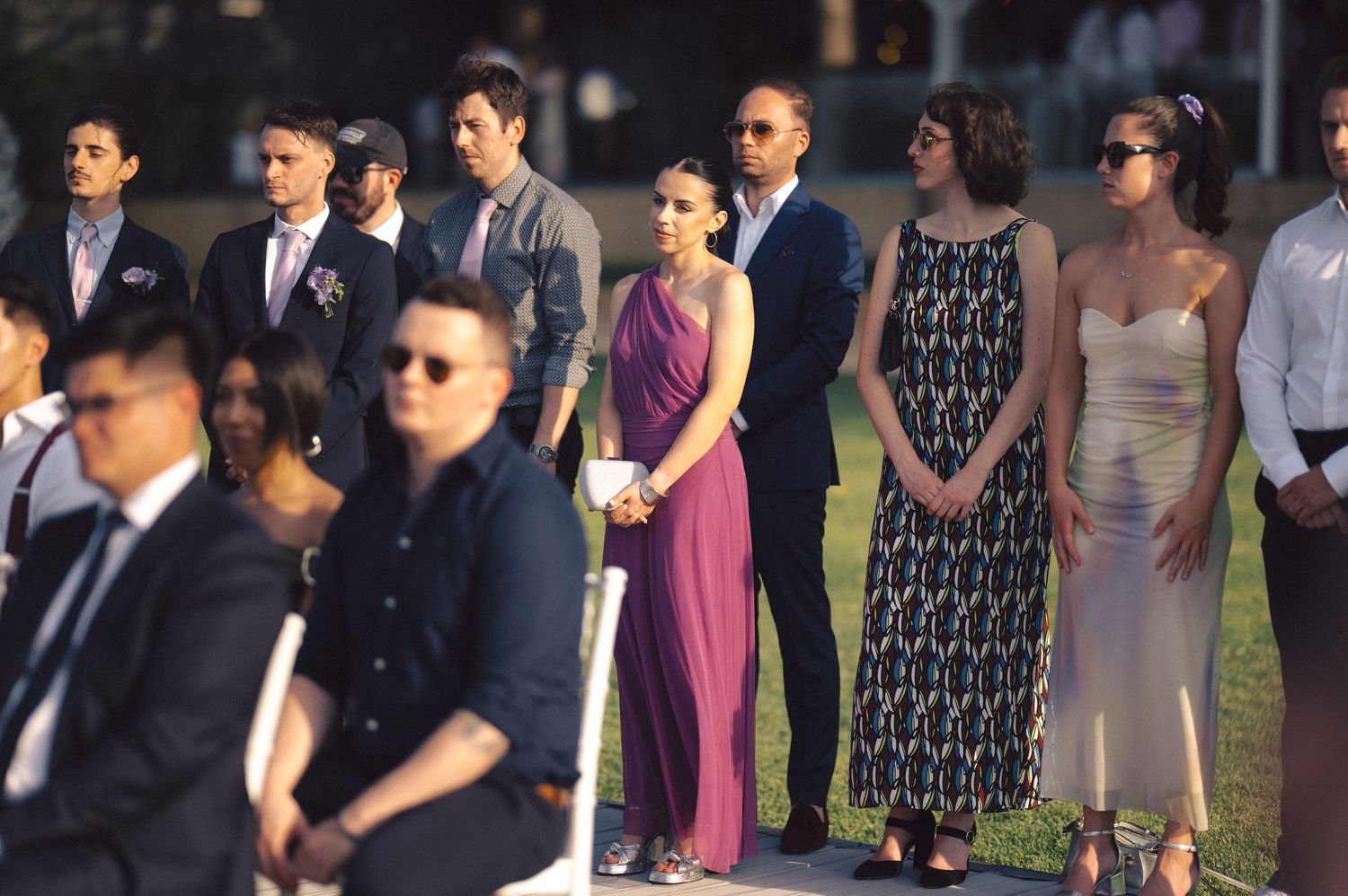 Well-dressed wedding guests stand in a line outdoors during an elegant summer ceremony.