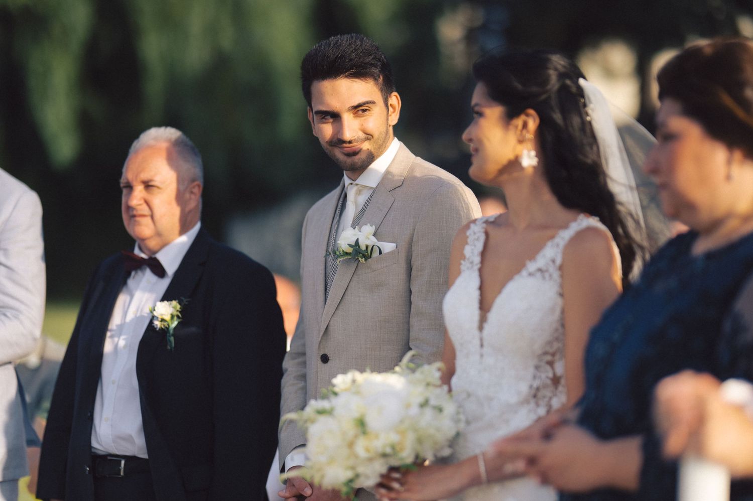 A wedding party stands together outdoors during a ceremony, with white floral bouquets and formal attire.