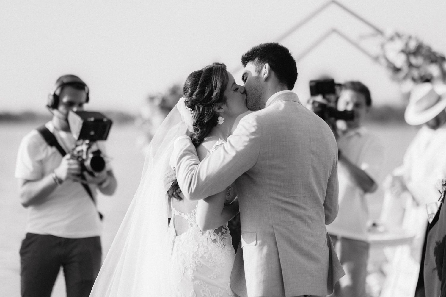Black and white photo of a couple sharing a romantic kiss at their outdoor wedding ceremony while photographer captures moment.