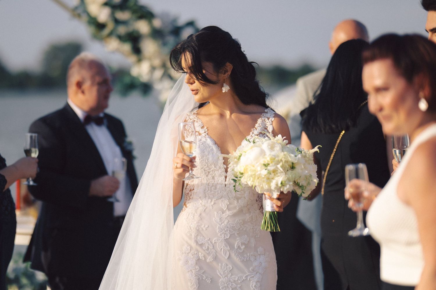 Wedding guests gather outdoors at an elegant reception as someone in a white lace gown holds a bouquet.