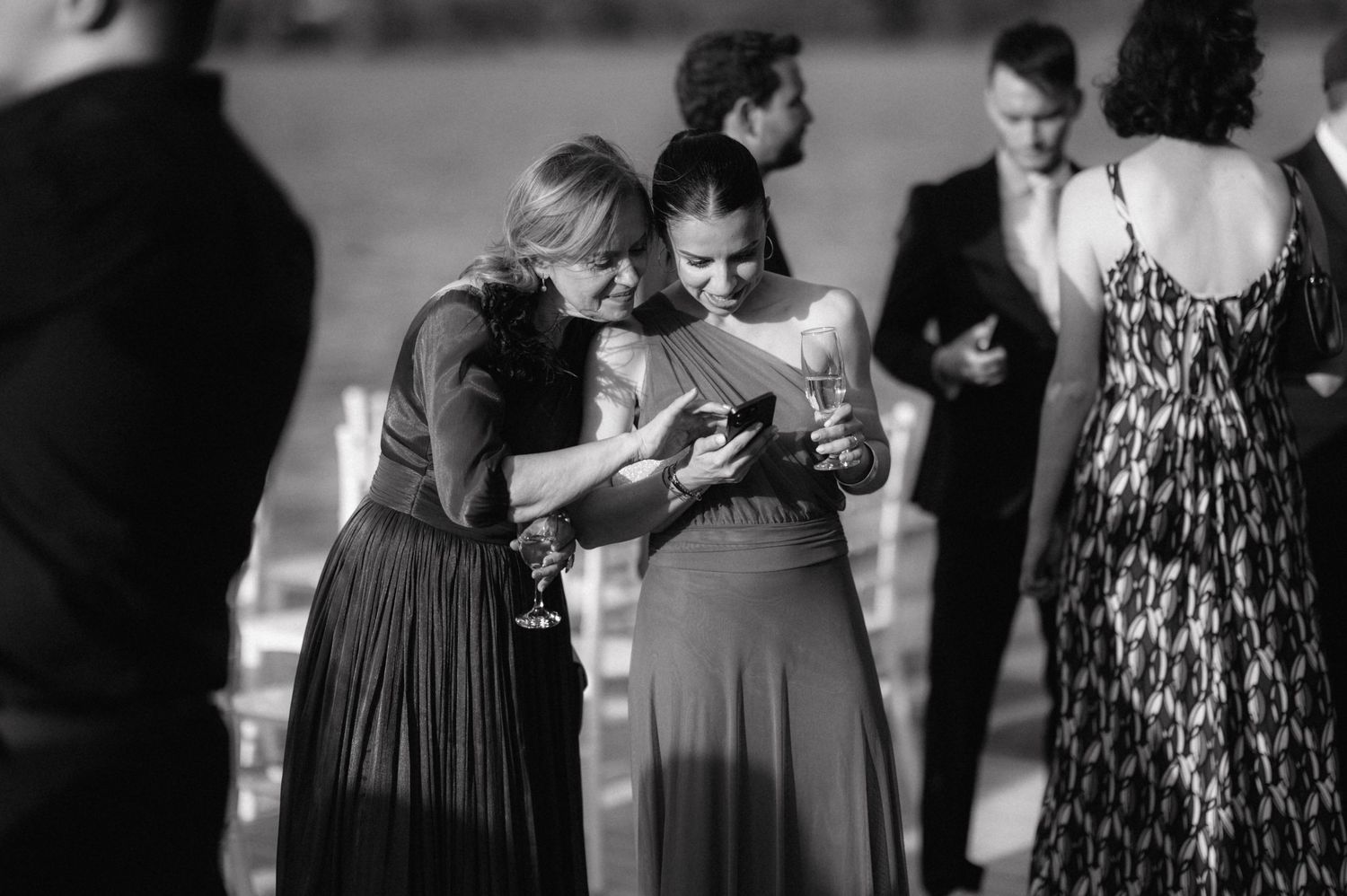 Two women in elegant dresses examine something on their phones during a formal event in black and white.