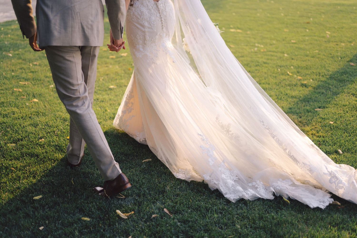 Close-up view of bride and groom walking on grass with white wedding dress train flowing behind.