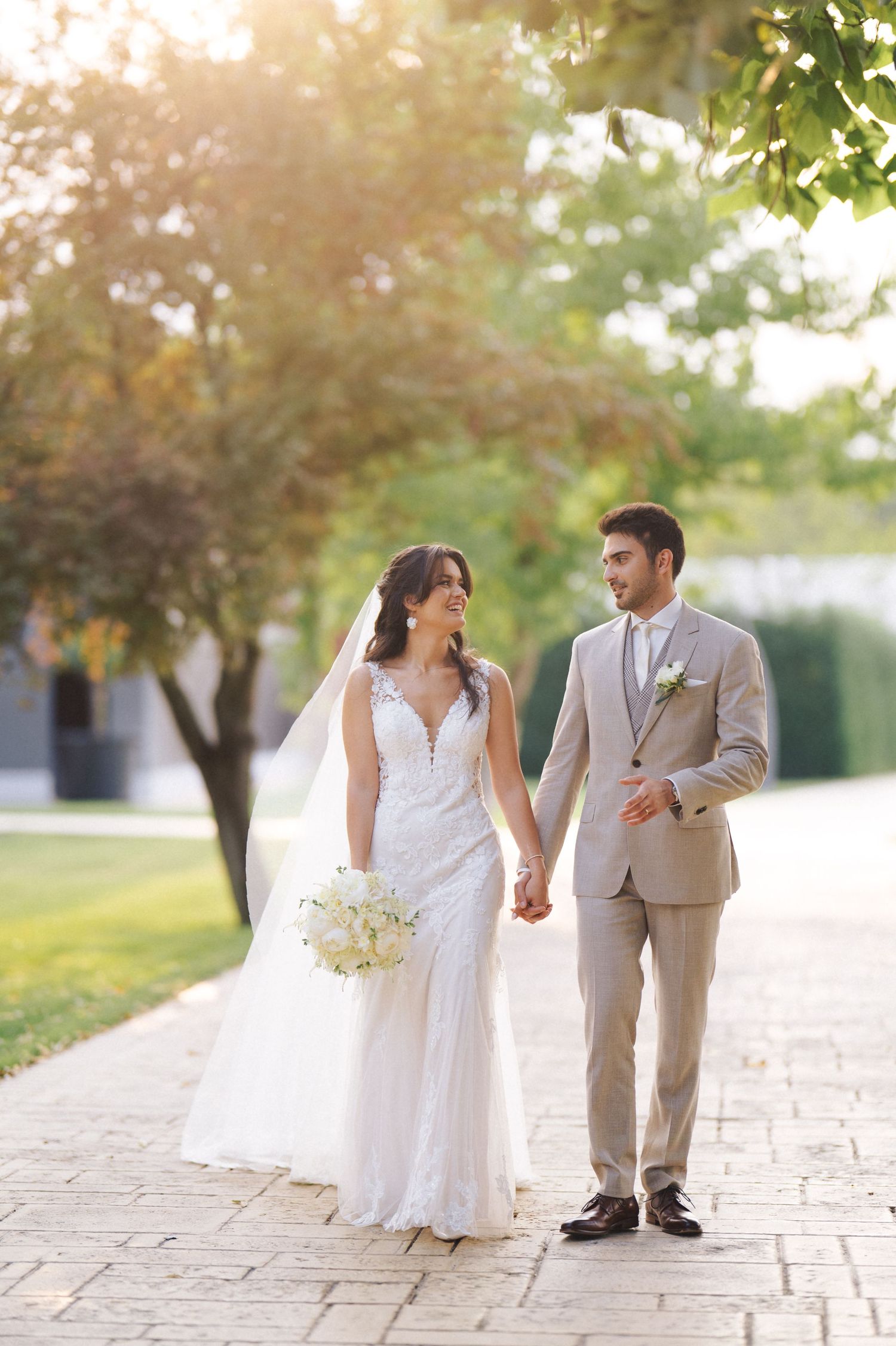 A couple walking hand-in-hand outdoors on stone walkway, with bride in white gown and groom in beige suit.