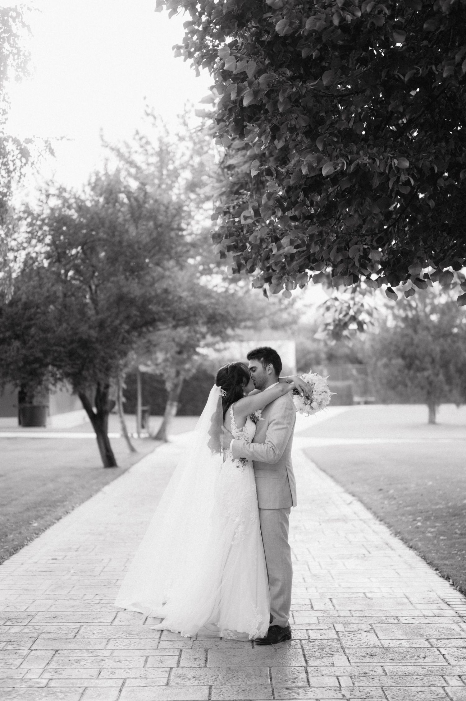 A romantic black and white wedding photo of a couple embracing on a tree-lined brick pathway.