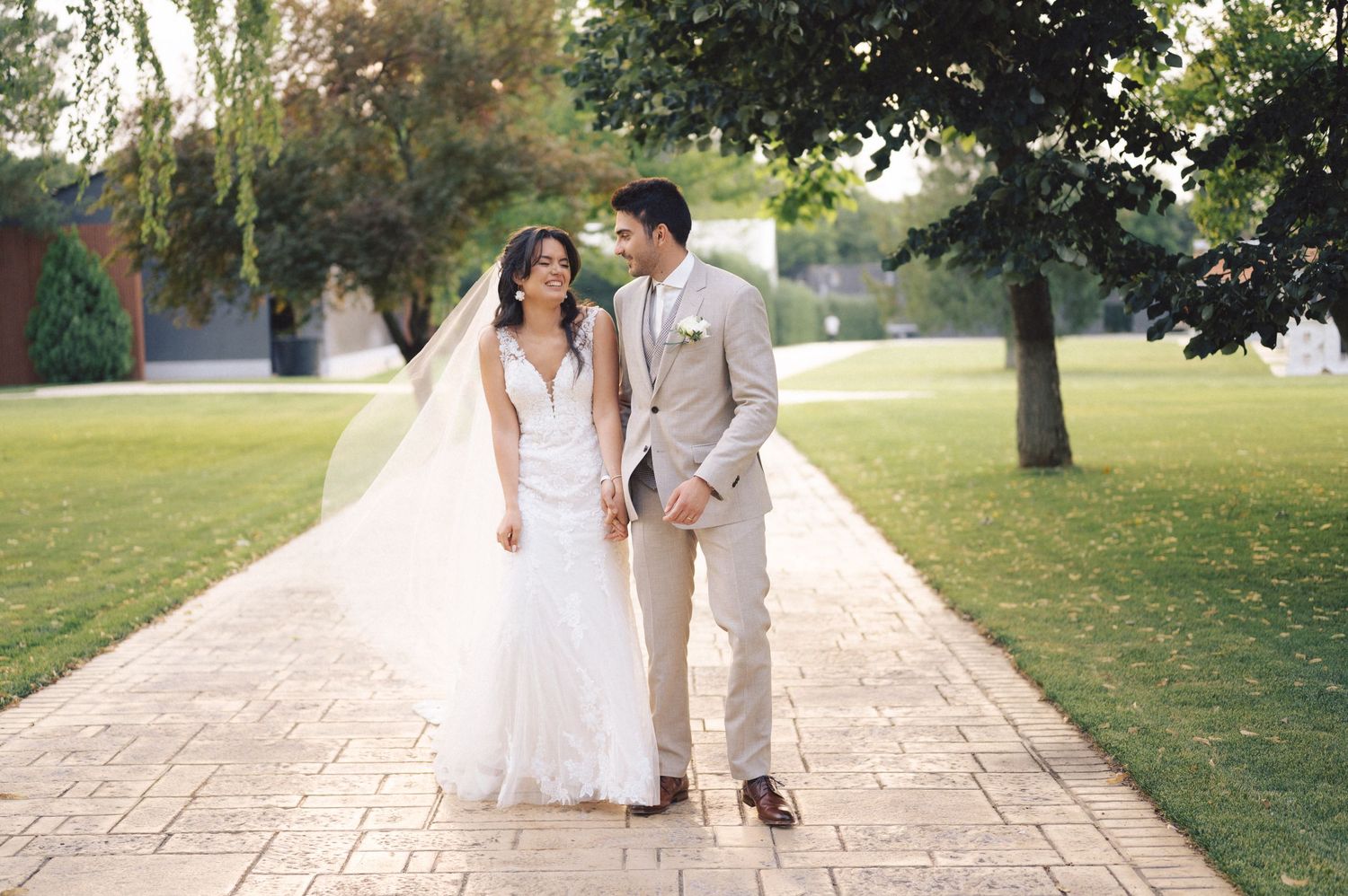 Newlyweds walking hand in hand down a paved garden path surrounded by green trees and lawns on a sunny day.
