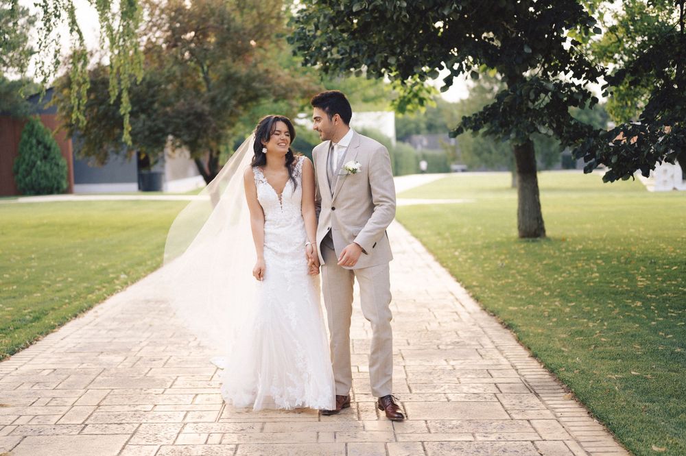 Newlyweds walking hand in hand down a paved garden path surrounded by green trees and lawns on a sunny day.