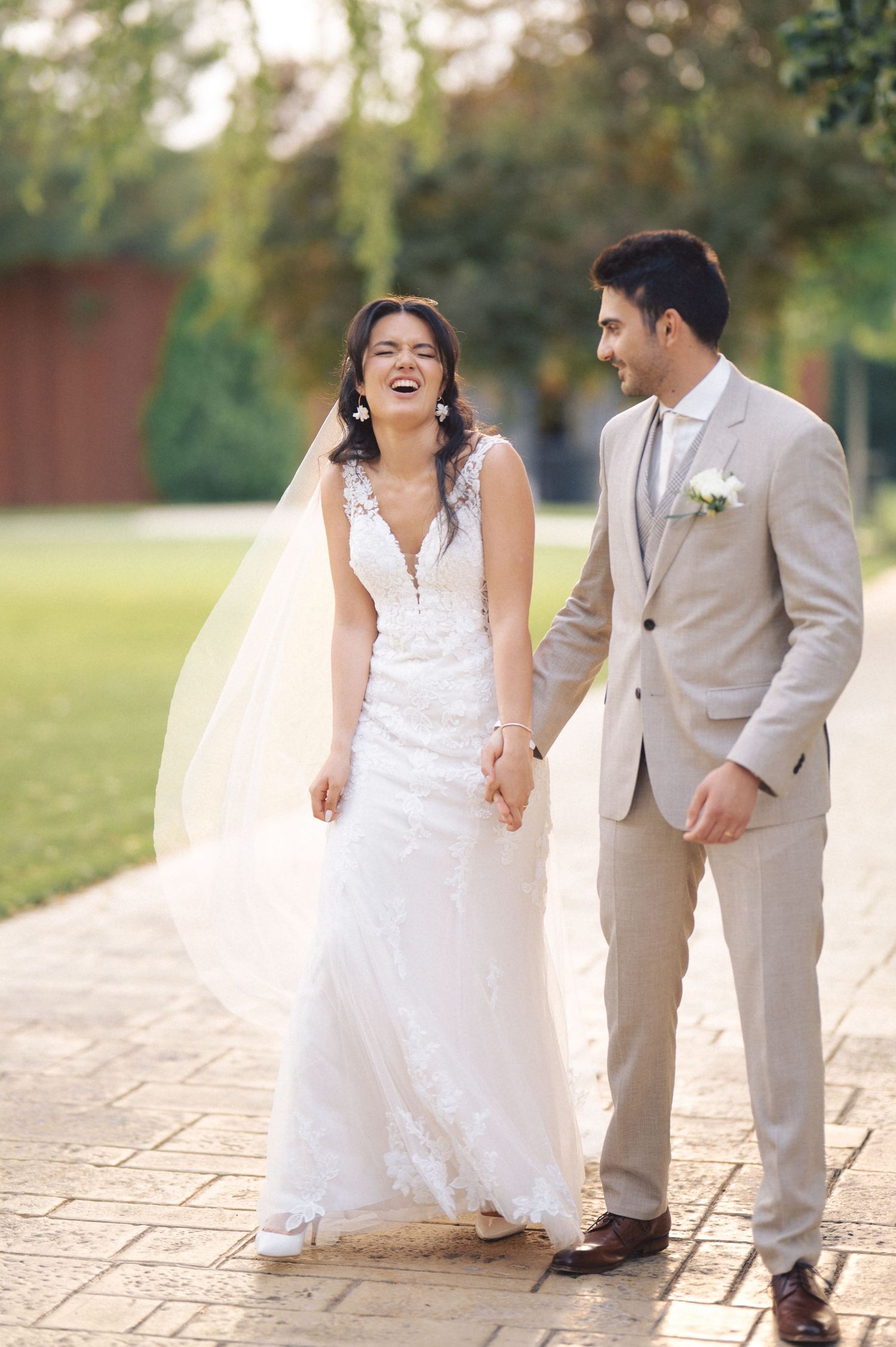 A happy couple on their wedding day walking outdoors in warm sunlight, wearing white lace gown and tan suit.