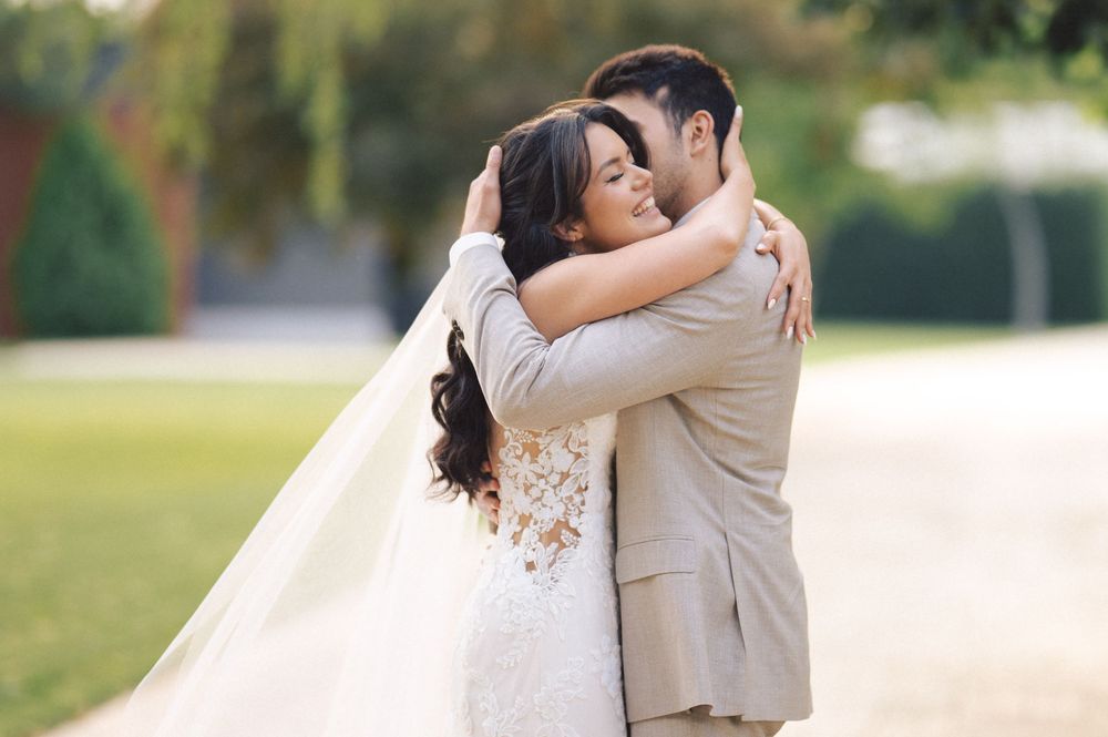 Newlyweds embrace outdoors on their wedding day, with bride in white lace gown and groom in light beige suit.