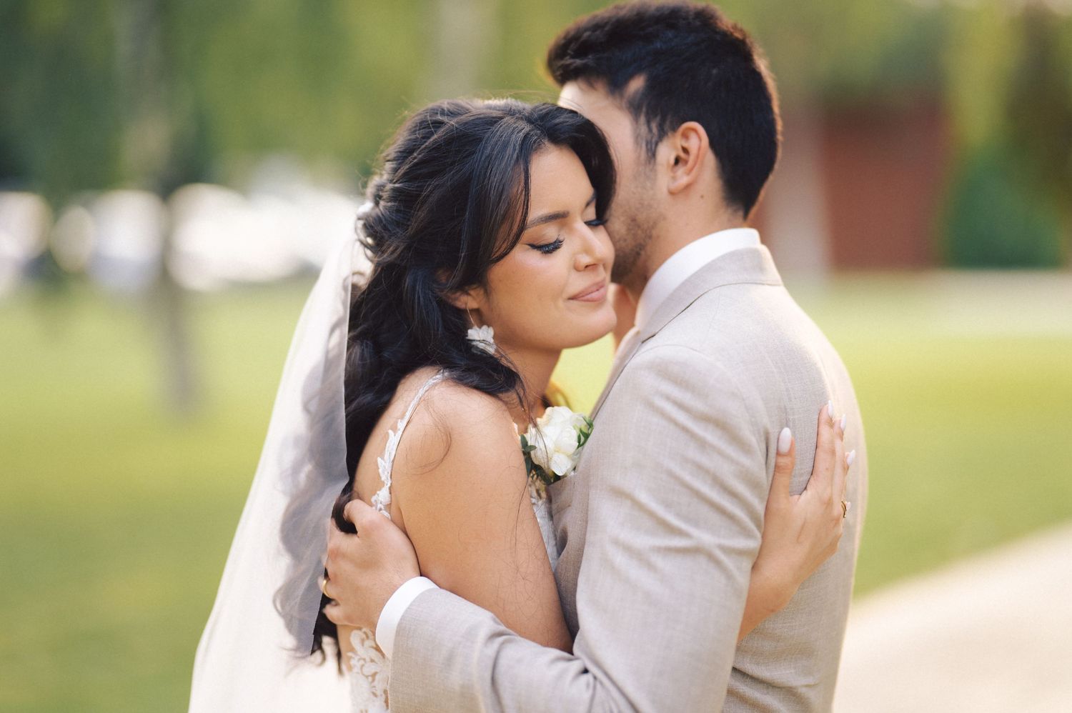 Newlywed couple embraces tenderly during outdoor wedding photoshoot with white bridal veil and soft natural lighting.