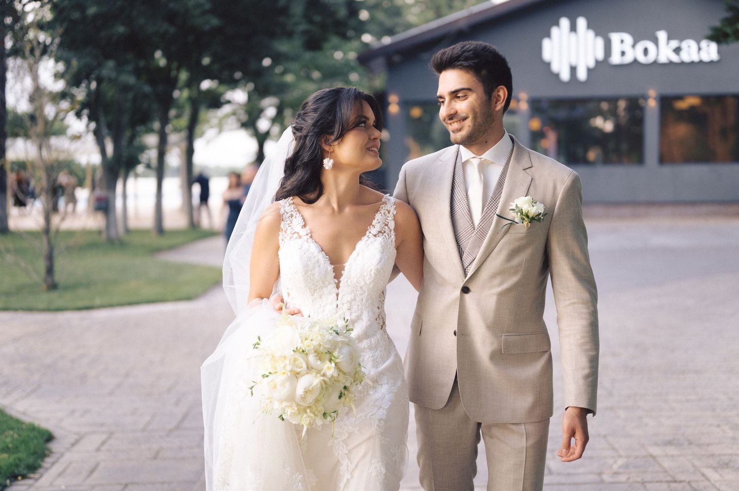 A bride in lace v-neck gown and groom in beige suit walk together outdoors while smiling.
