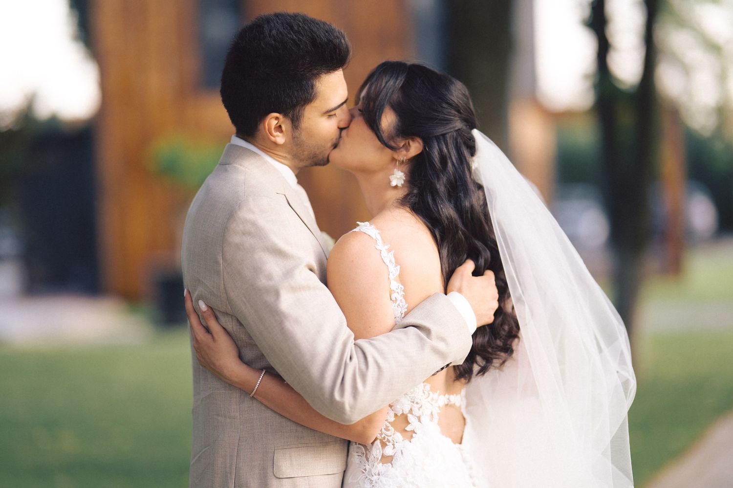 A newlywed couple shares a romantic kiss outdoors while wearing traditional white wedding attire.