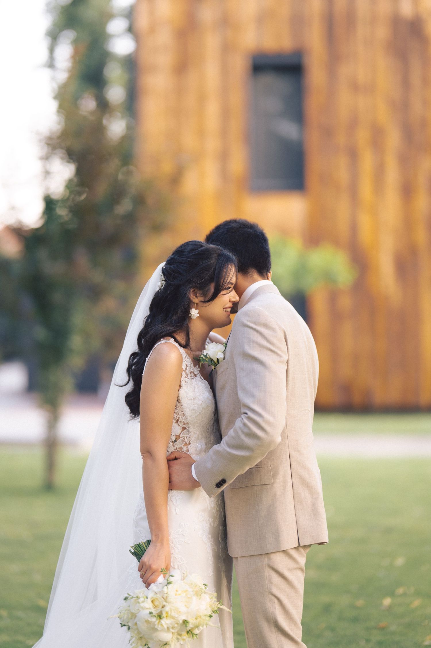 A bride in a white lace dress and groom in a beige suit share an intimate moment at a rustic barn wedding venue.