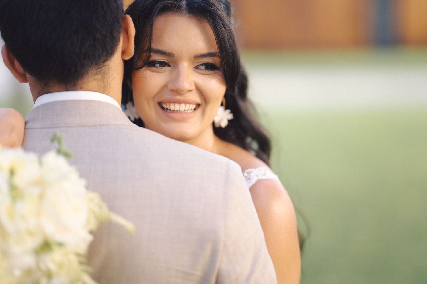 Smiling bride in white dress looks over shoulder of groom in beige suit during outdoor wedding ceremony.