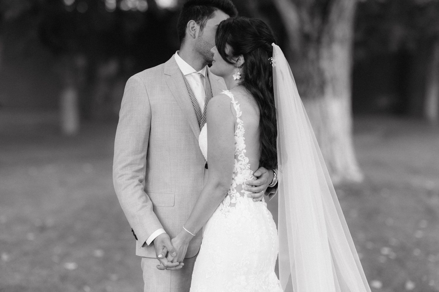 A black and white photograph of newlyweds sharing a tender moment outdoors with bride in lace gown and flowing veil.