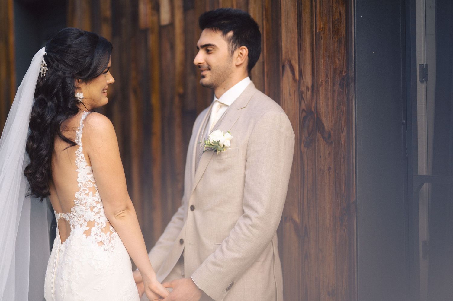 Elegant rustic wedding scene with couple by wooden wall in matching cream and white attire.