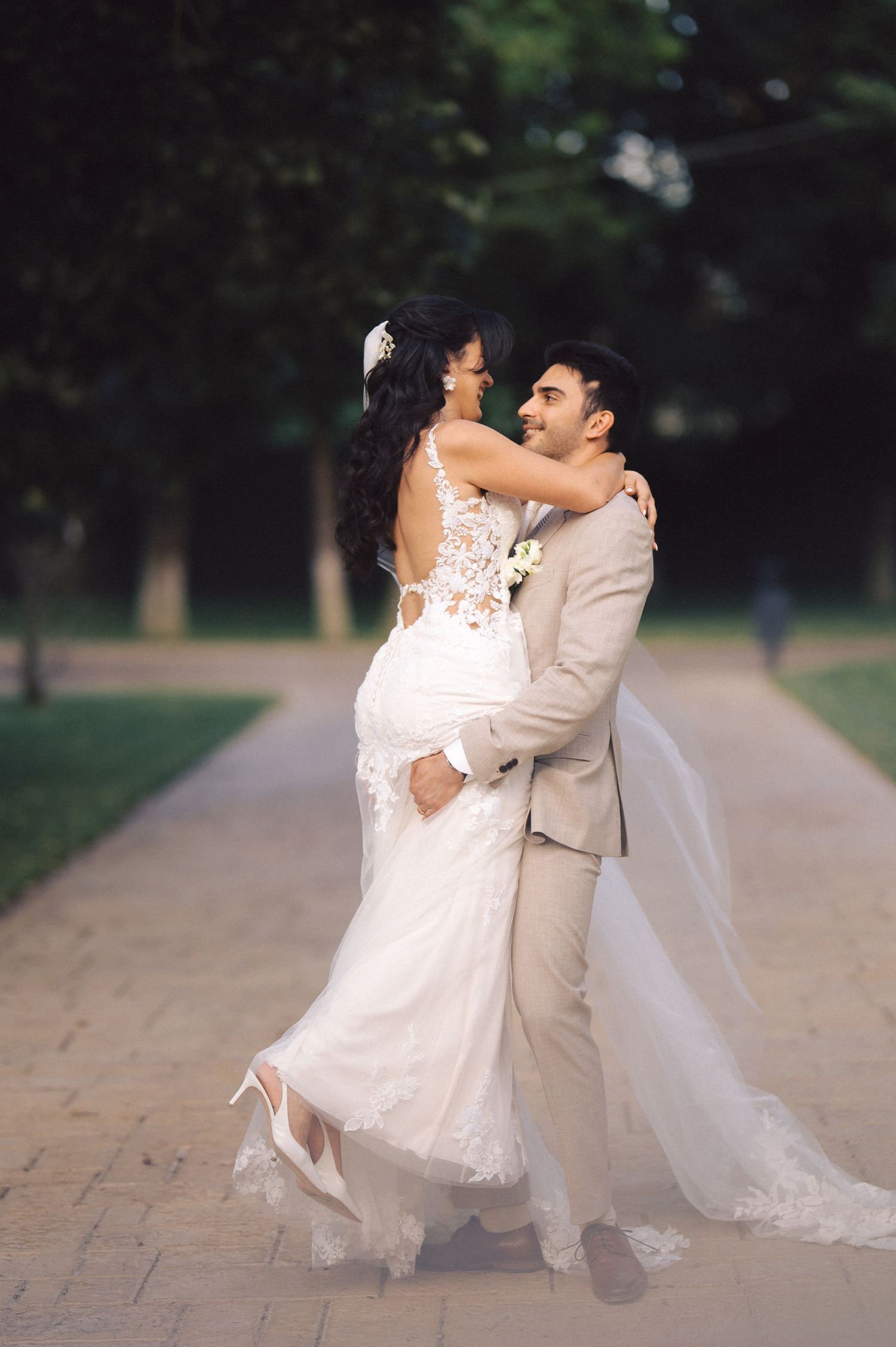 A romantic wedding couple embraces on a tree-lined path, with the bride wearing a lace white gown and groom in beige suit.