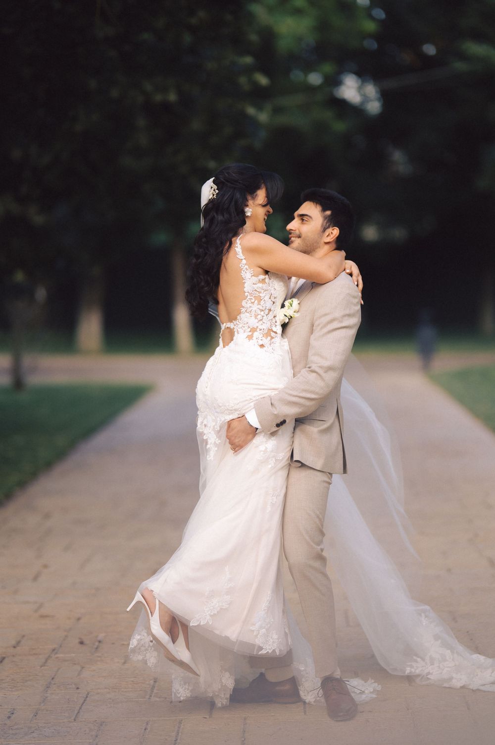 A romantic wedding couple embraces on a tree-lined path, with the bride wearing a lace white gown and groom in beige suit.