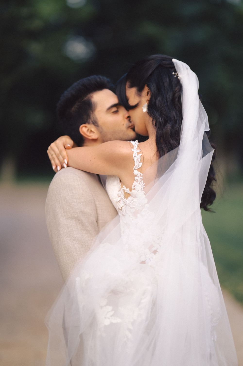 A romantic wedding photo captured outdoors featuring a couple sharing a kiss, with the bride wearing a white veil and lace dress.