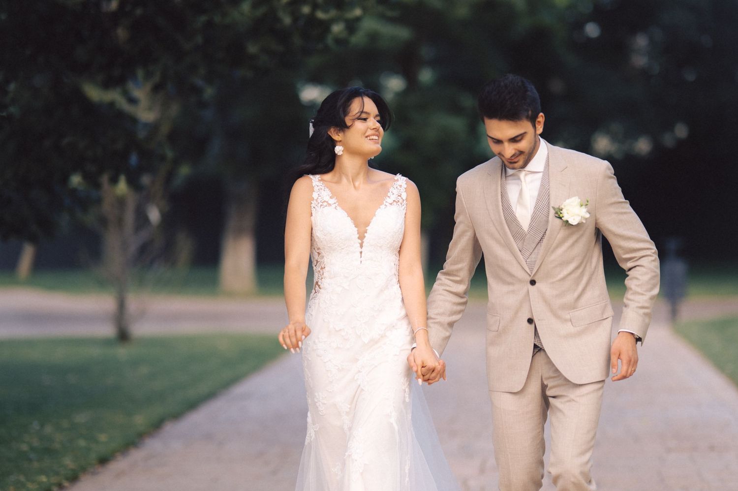 A couple wearing white and beige outfits walk hand in hand along a garden pathway during a romantic outdoor photo shoot.