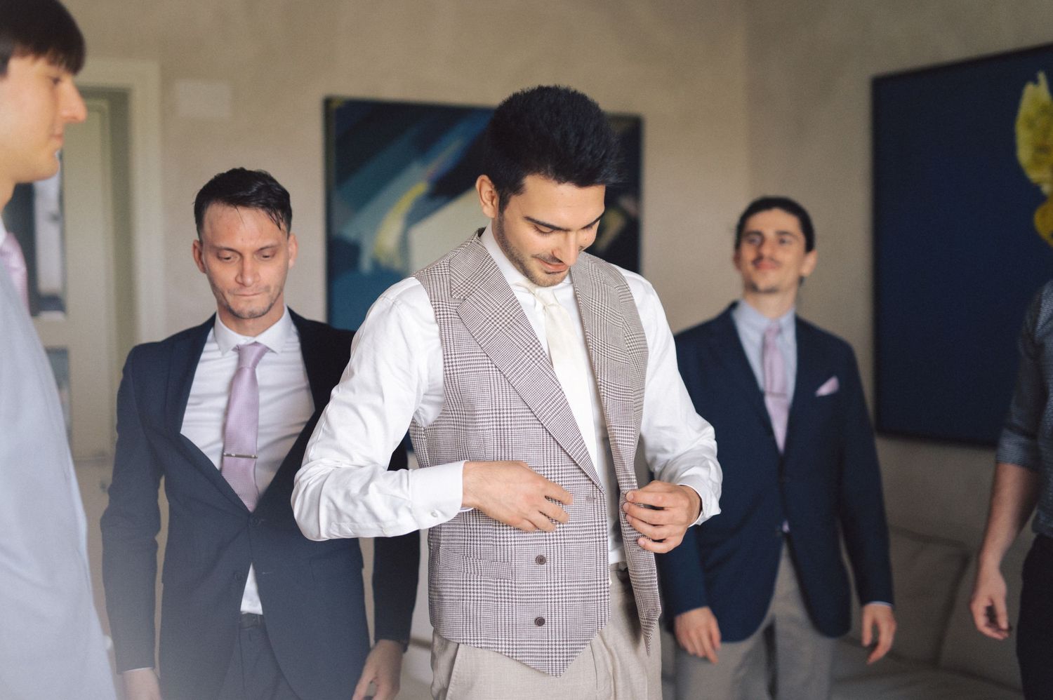 A group of well-dressed men in suits and formal attire prepare for a special event or wedding in a hallway.
