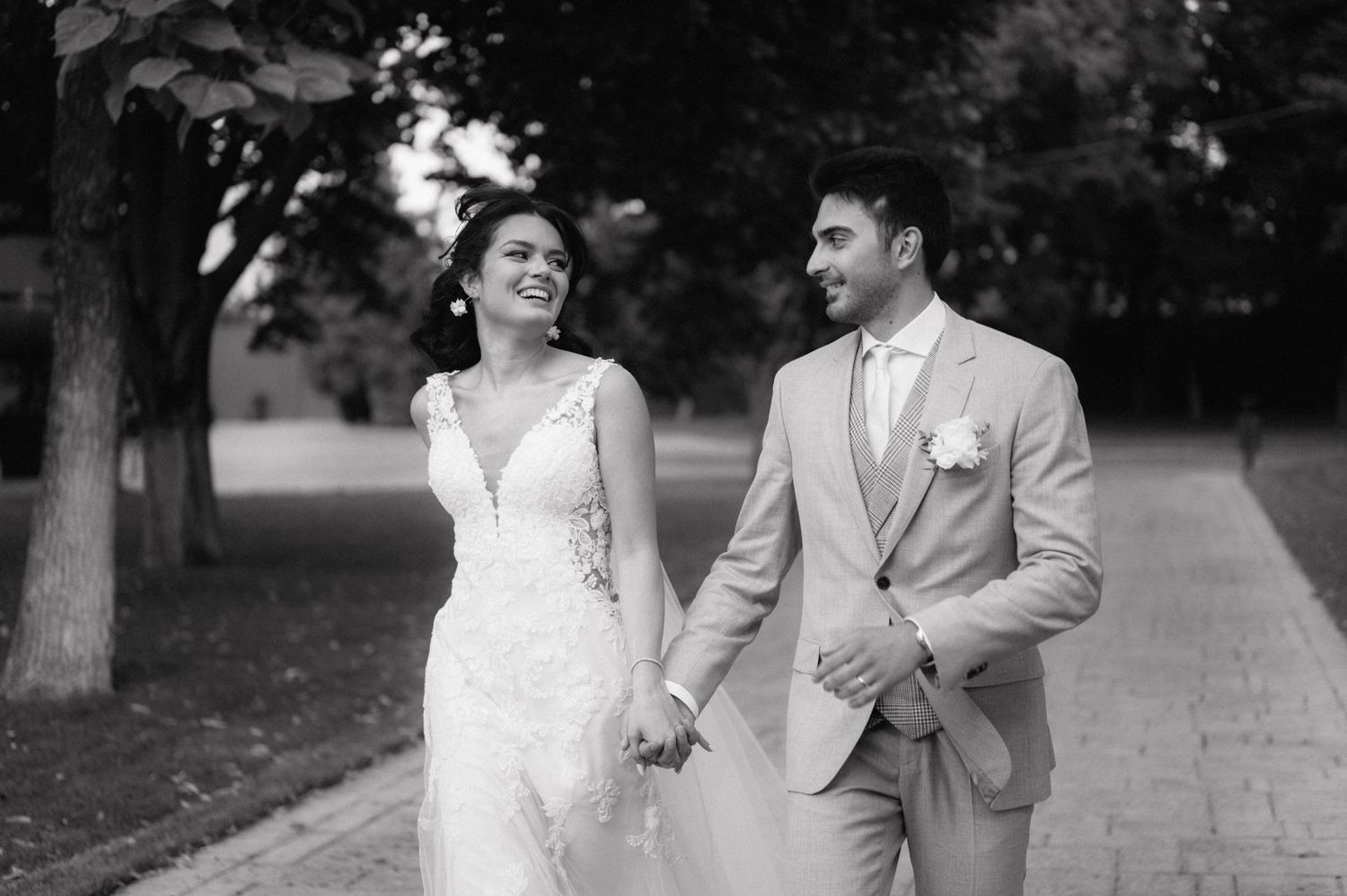 A couple walking and laughing together outdoors in formal attire on a tree-lined path.