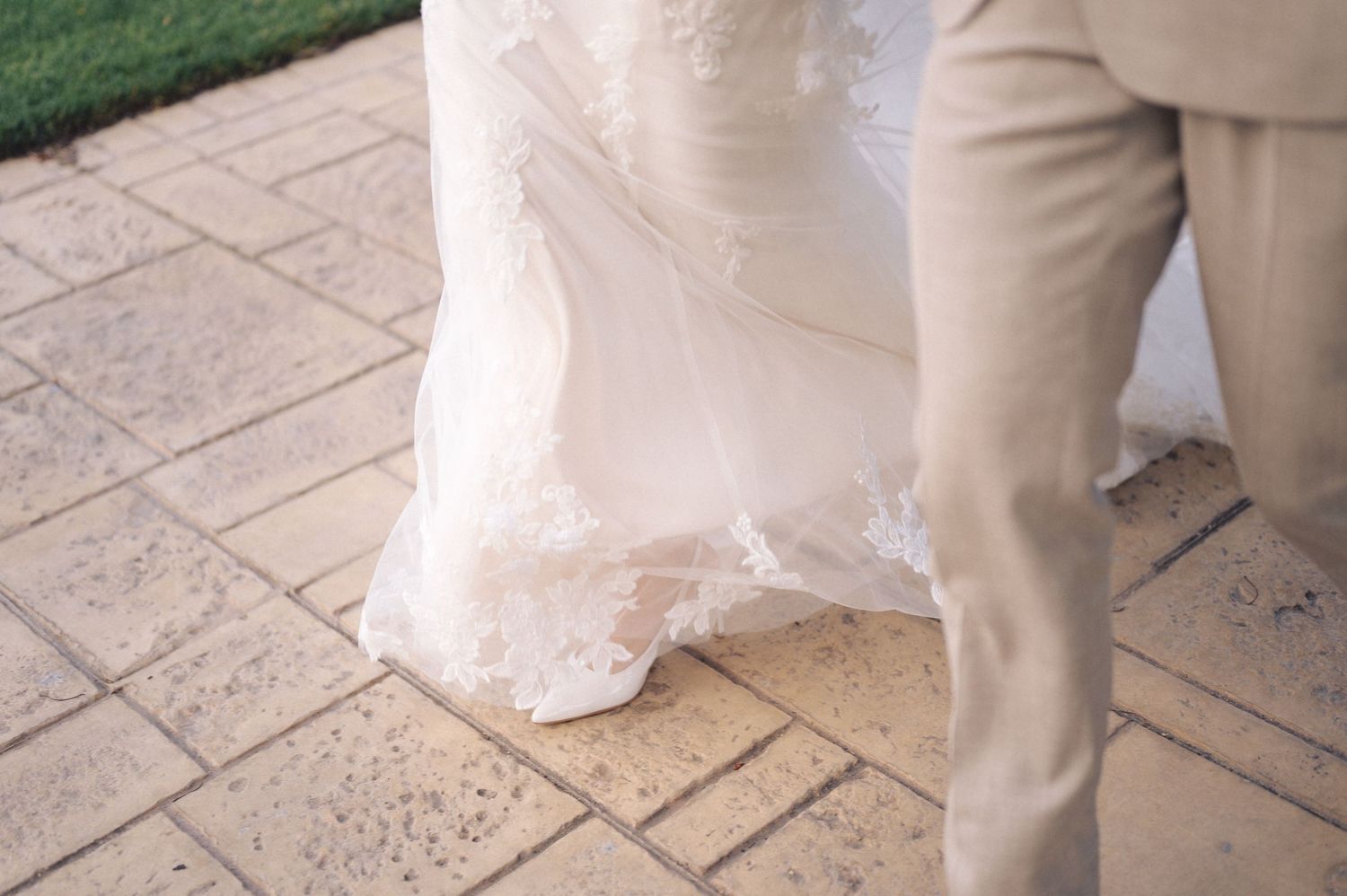 Bottom portion of a white wedding dress and beige suit pants standing on patterned stone pavers.