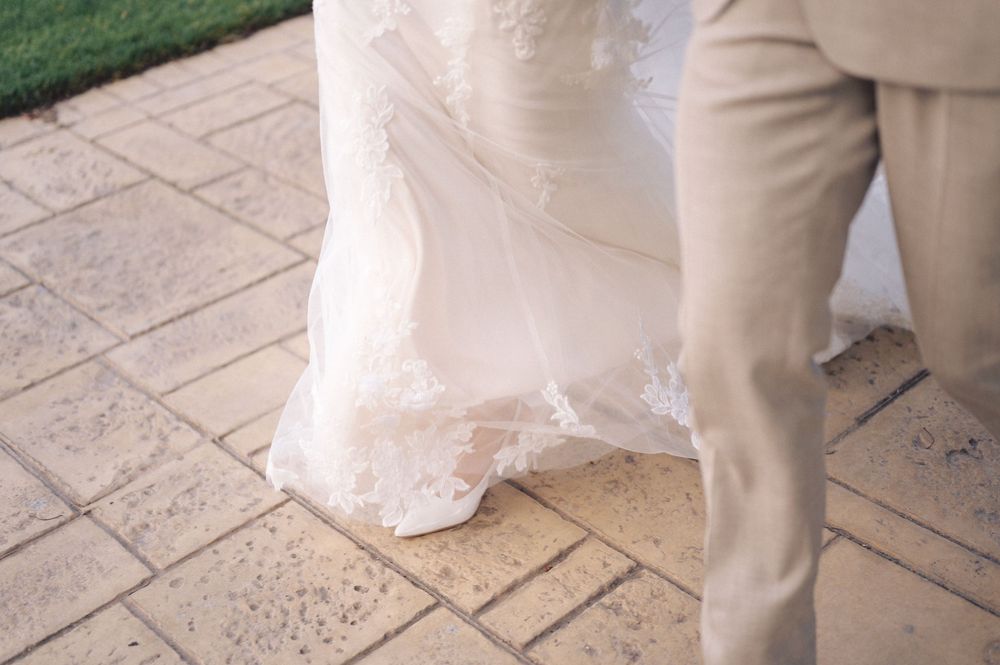 Bottom portion of a white wedding dress and beige suit pants standing on patterned stone pavers.