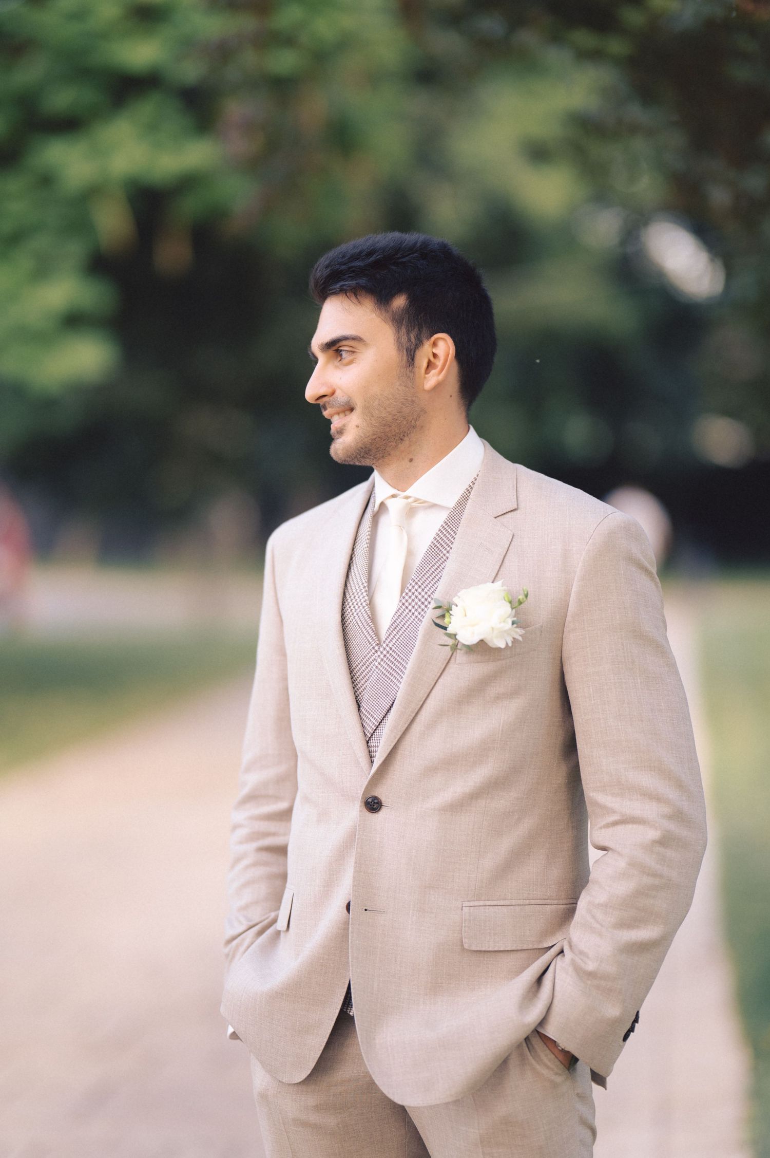 A groom in a light beige suit and white boutonniere poses outdoors with a natural blurred background.