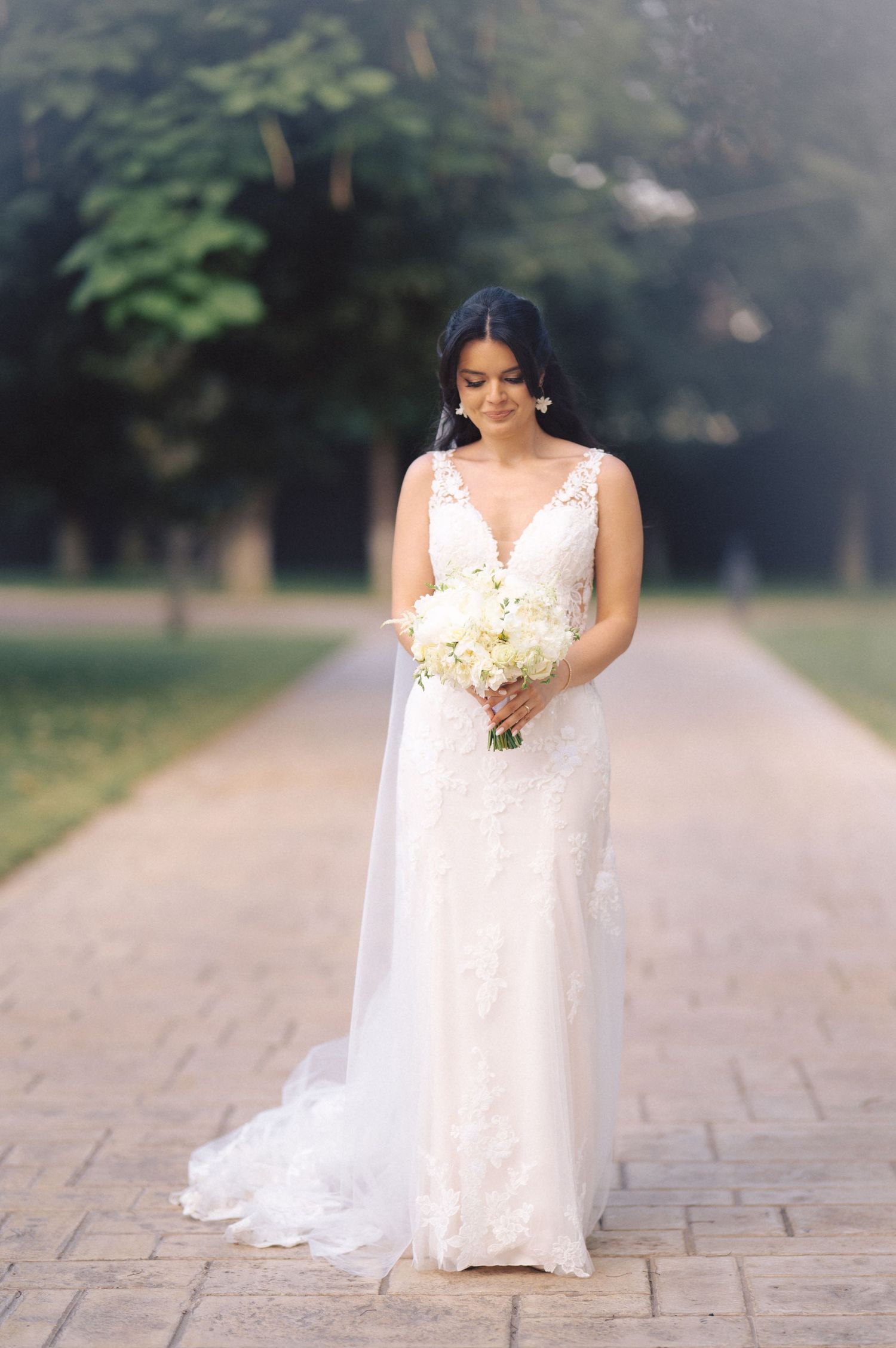 A bride in an elegant white lace wedding dress poses outdoors on a tree-lined stone pathway.