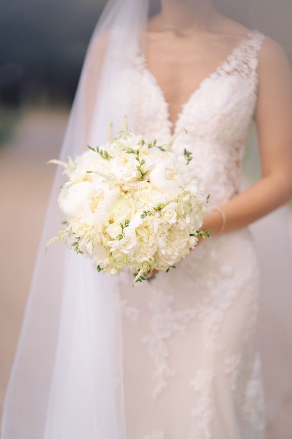 Elegant white bridal bouquet with peonies and delicate flowers shown with lace wedding dress and flowing veil.
