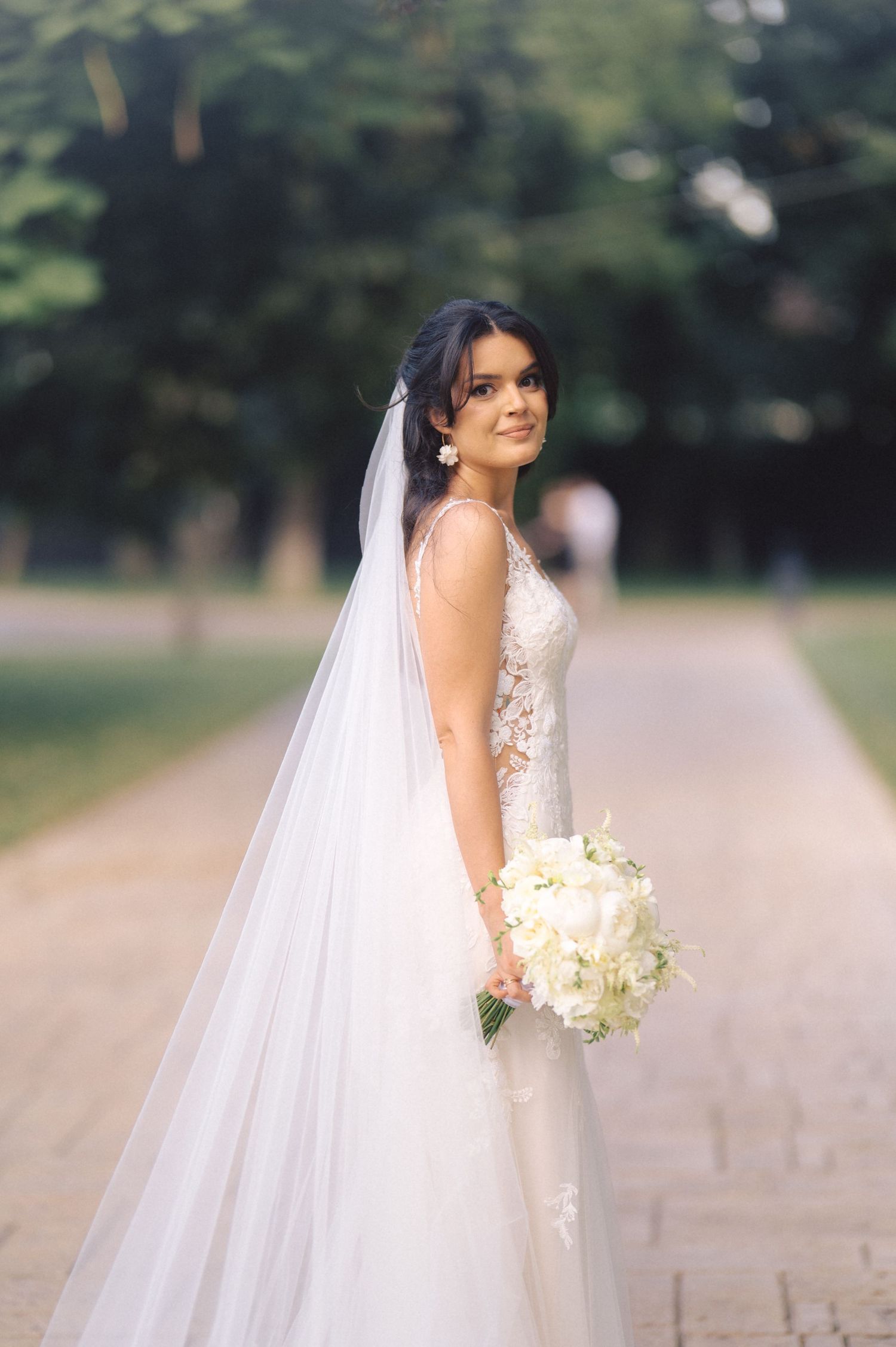 A bride wearing a white lace wedding dress and long veil holds a white bouquet while posing on a tree-lined path.