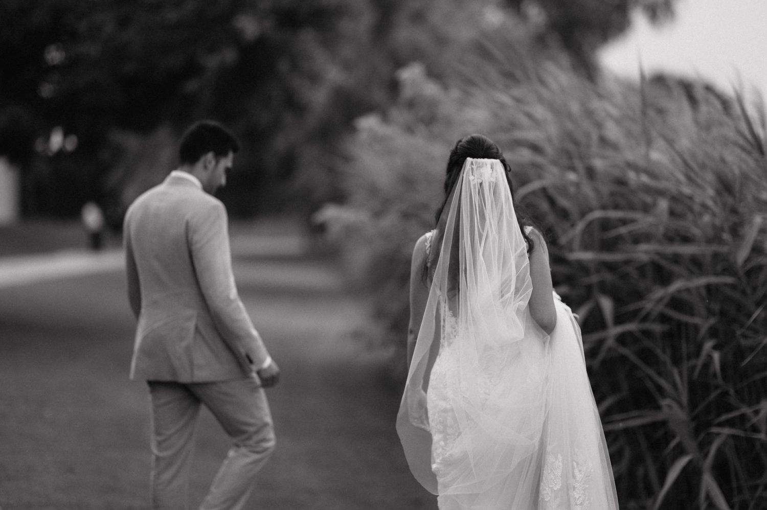 A couple in wedding attire walks down a path surrounded by lush green gardens at a beautiful outdoor venue.