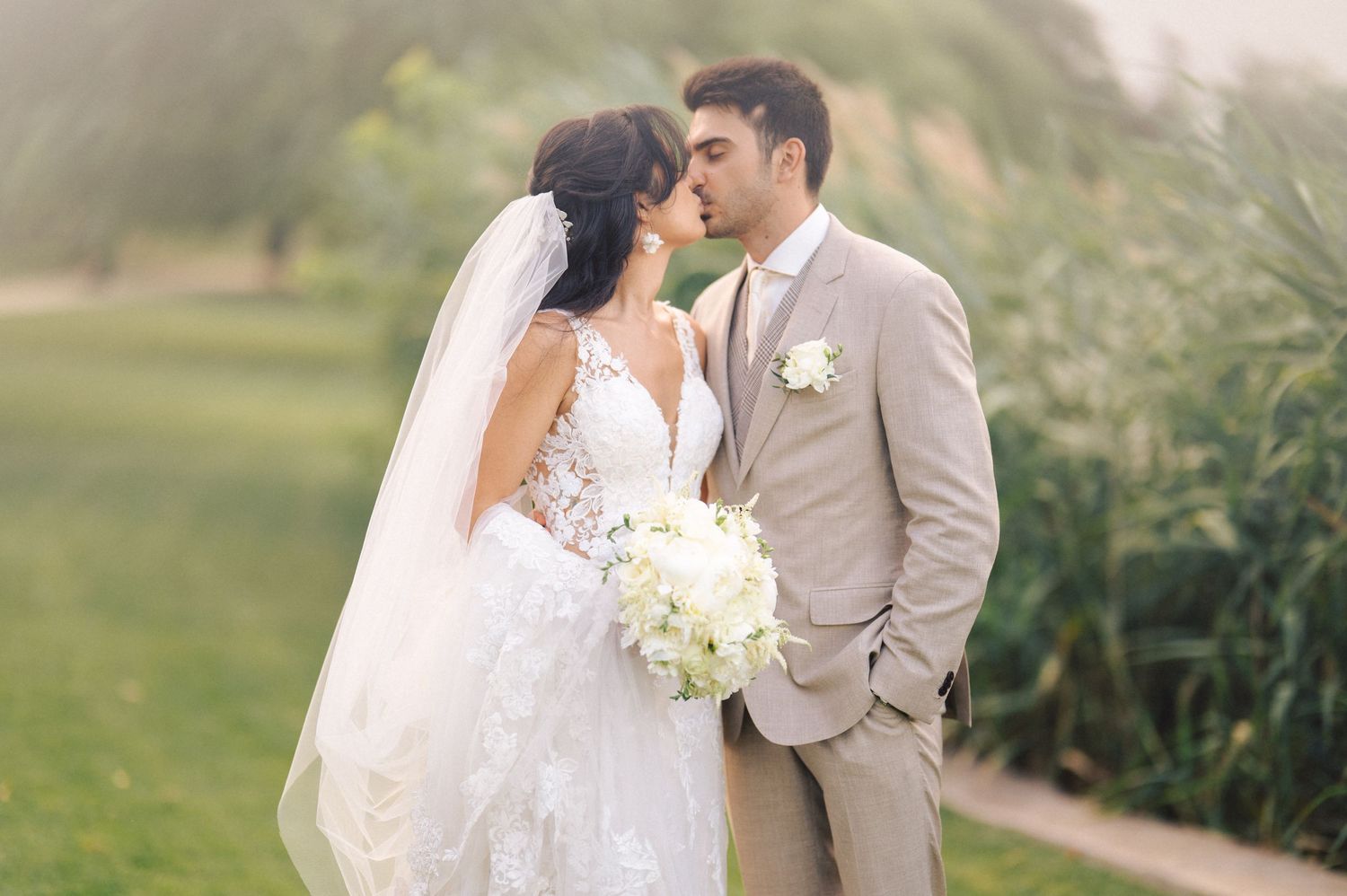 Newlyweds share a romantic moment in a garden setting, with the bride holding a white bouquet wearing a flowing veil.
