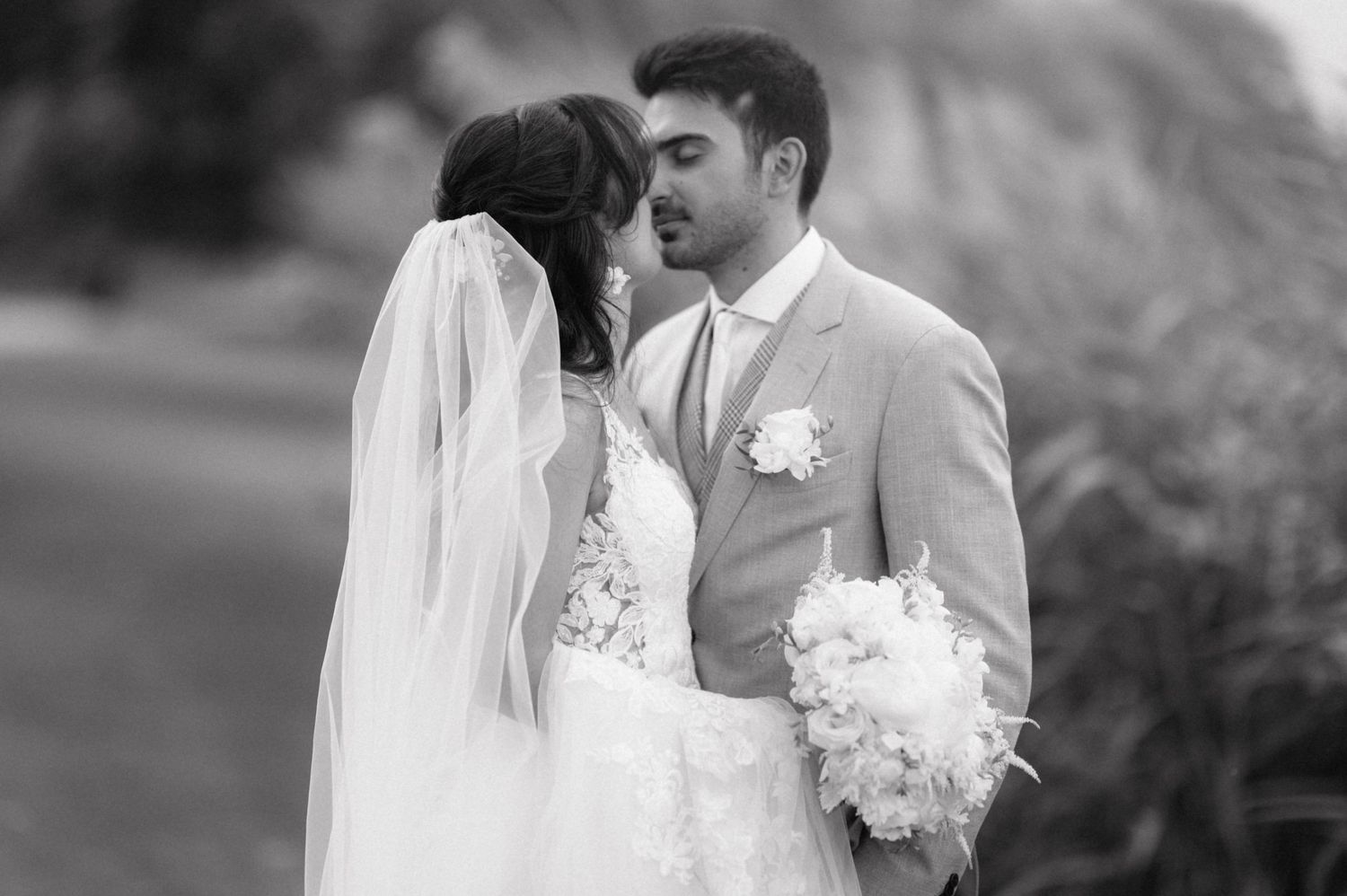 A romantic black and white wedding photo showing a tender moment between newlyweds with a flowing veil and bouquet.
