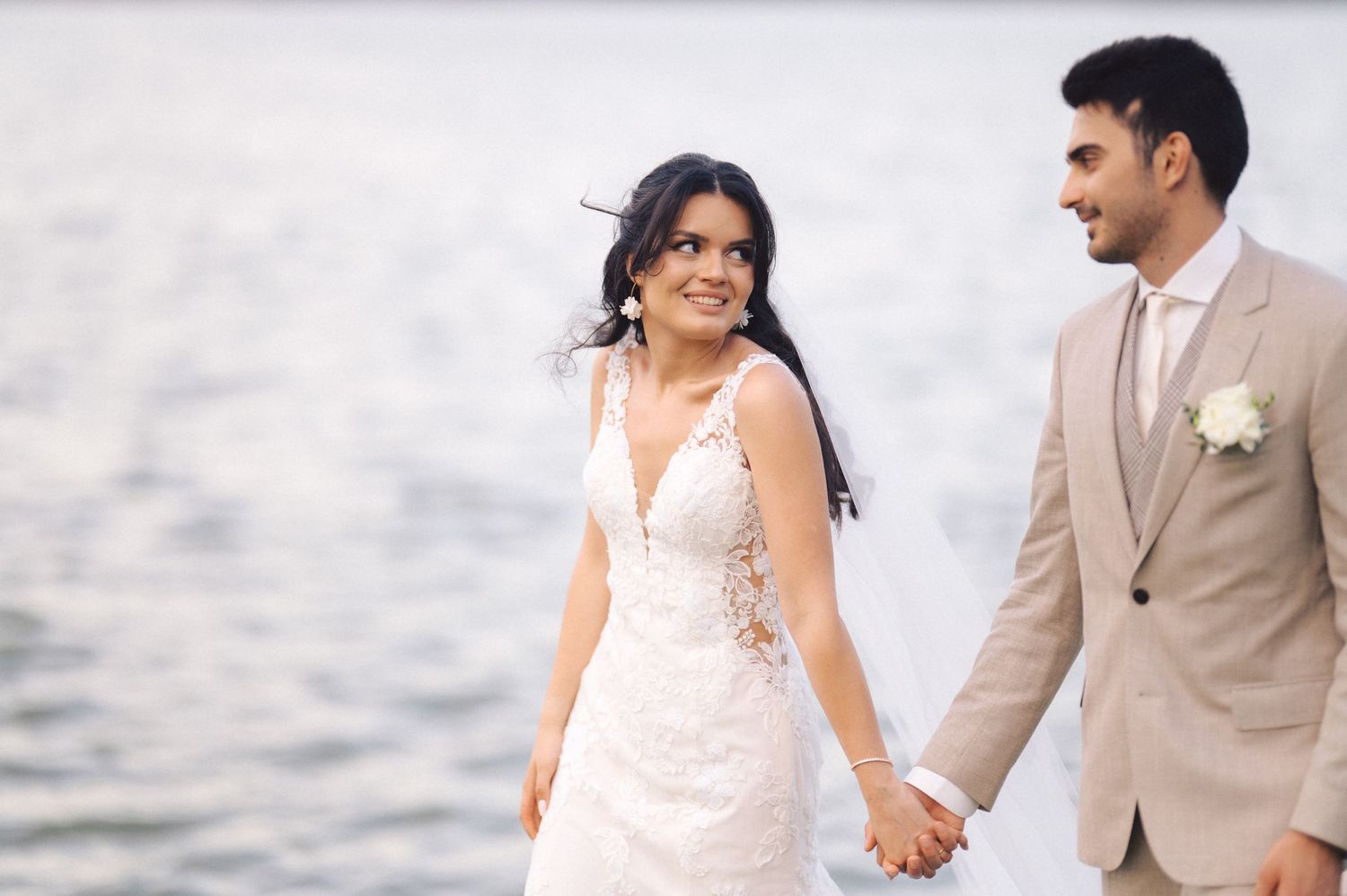Couple holding hands and walking on sandy beach in formal wedding attire during sunset.